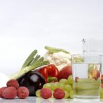 Un bicchiere d'acqua su un tavolo con frutta fresca sullo sfondo, simbolo di idratazione e salute.